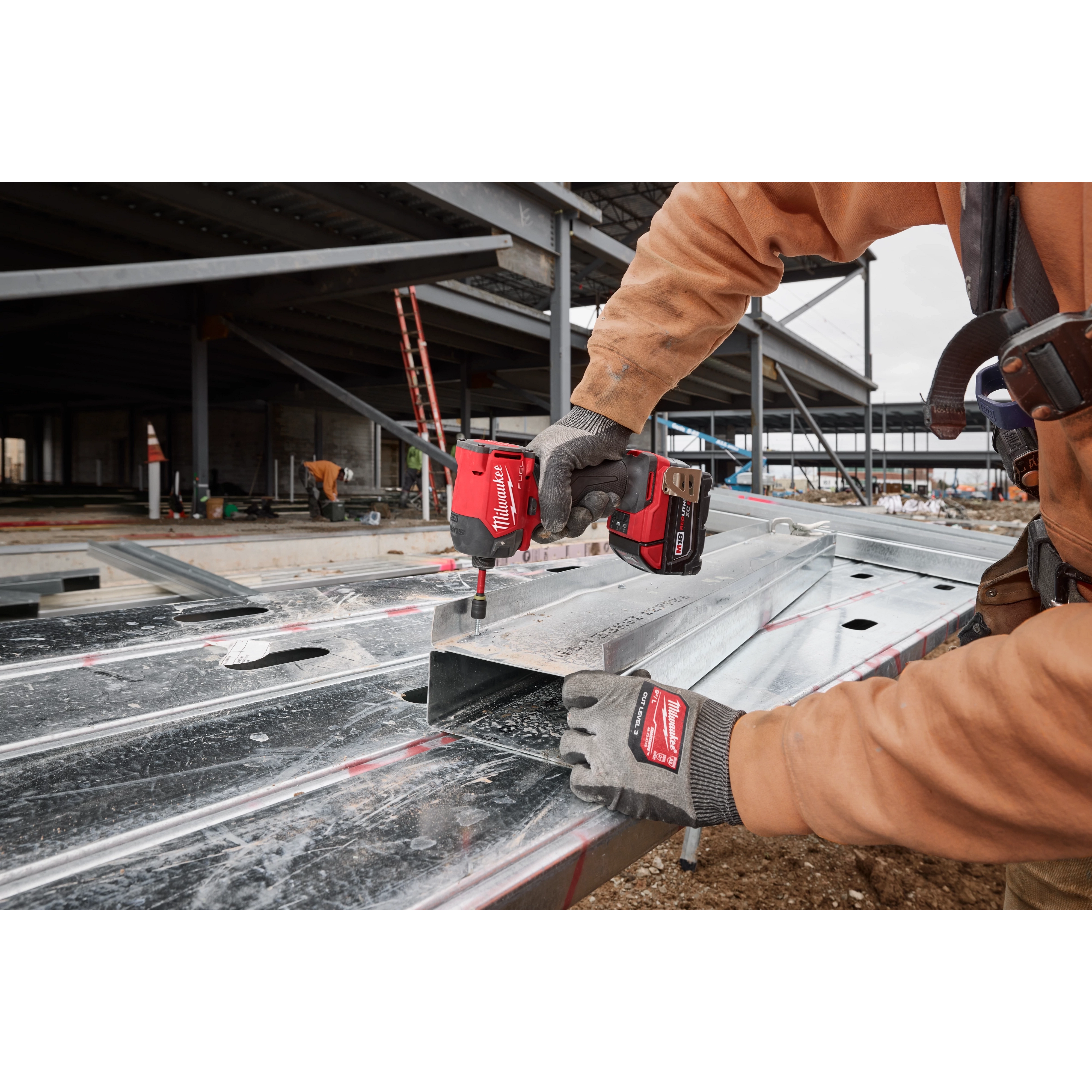 A person uses the M18 FUEL™ SURGE™ 1/4" Hex Hydraulic Driver to secure a metal sheet at a construction site. The tool is red and black, and the user is wearing gloves and a brown work jacket. Construction materials and partially built structures are visible in the background.