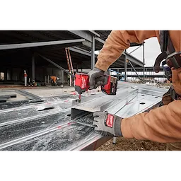 A person uses the M18 FUEL™ SURGE™ 1/4" Hex Hydraulic Driver to secure a metal sheet at a construction site. The tool is red and black, and the user is wearing gloves and a brown work jacket. Construction materials and partially built structures are visible in the background.