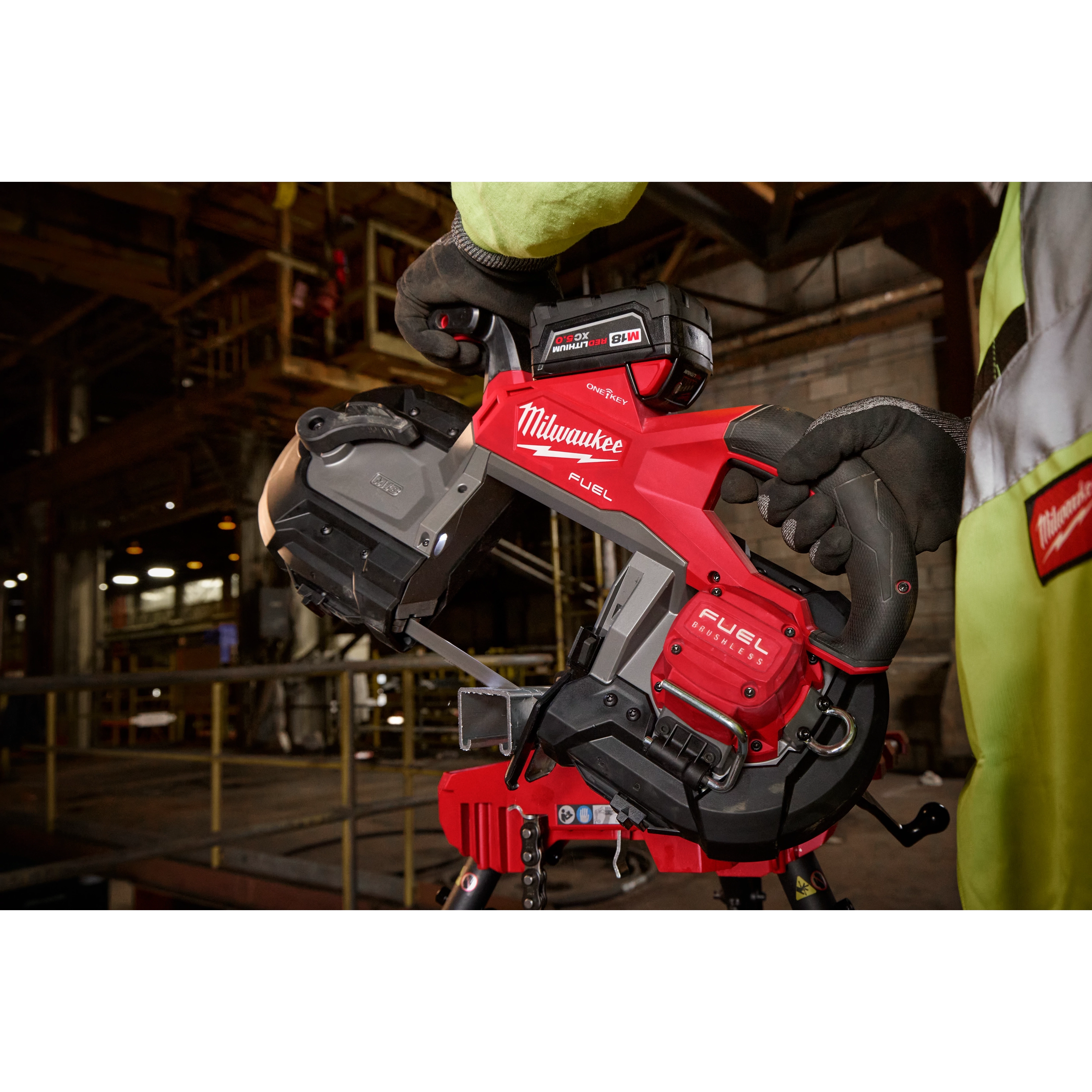 A worker wearing gloves holds a red and black M18 FUEL™ Deep Cut Dual Trigger Band Saw w/ ONE-KEY™ in an industrial setting. The tool features a visible battery pack and branding.