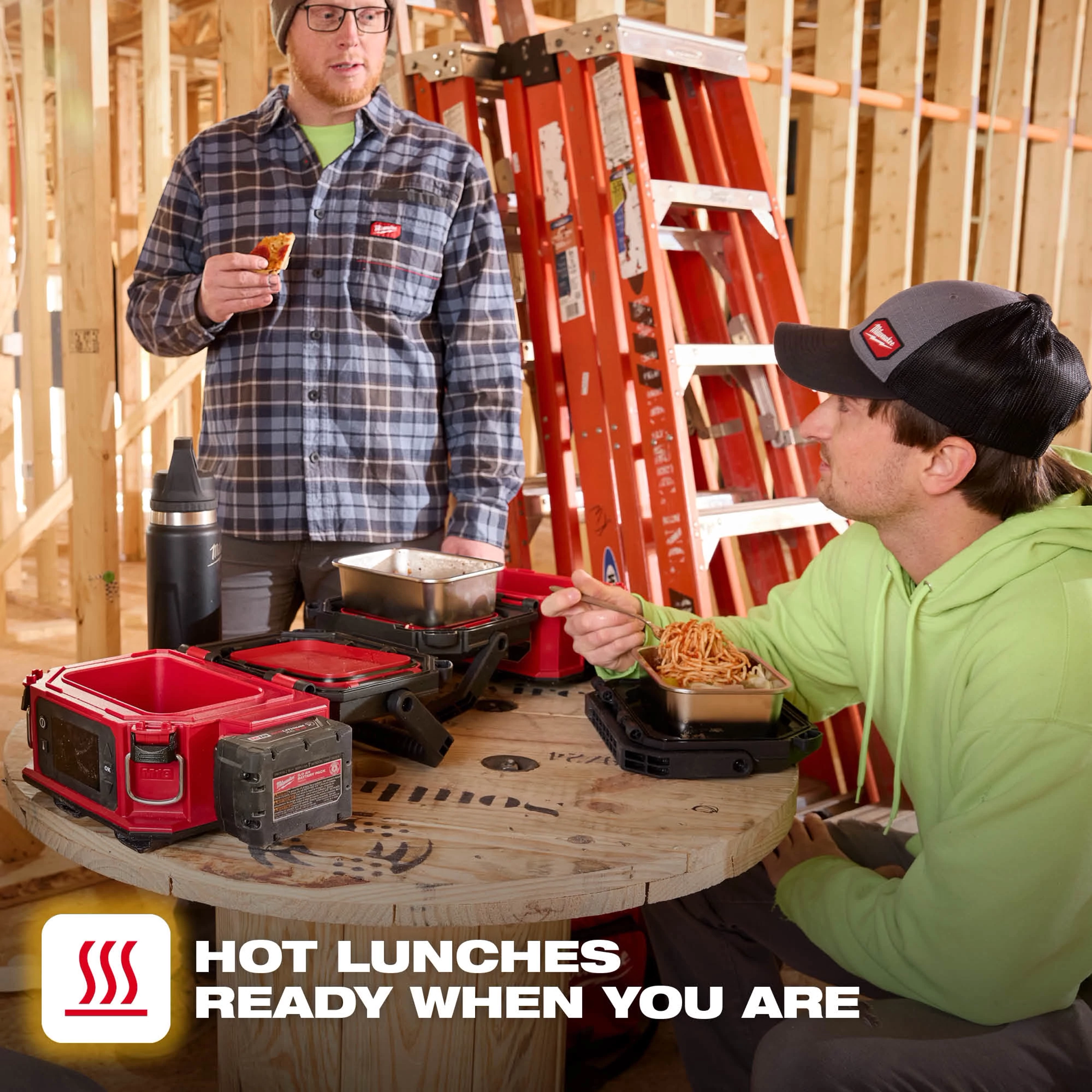 Workers eating at a wooden spool table with an open M18 Jobsite Lunch Warmer on a construction site.