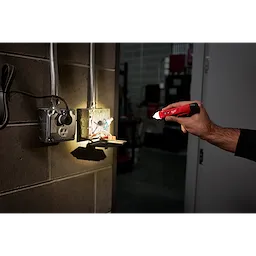 A person uses a Voltage Detector w/ LED to test an open electrical outlet box in a dimly lit room. The detector's LED light is illuminated, highlighting the wiring inside the electrical box.