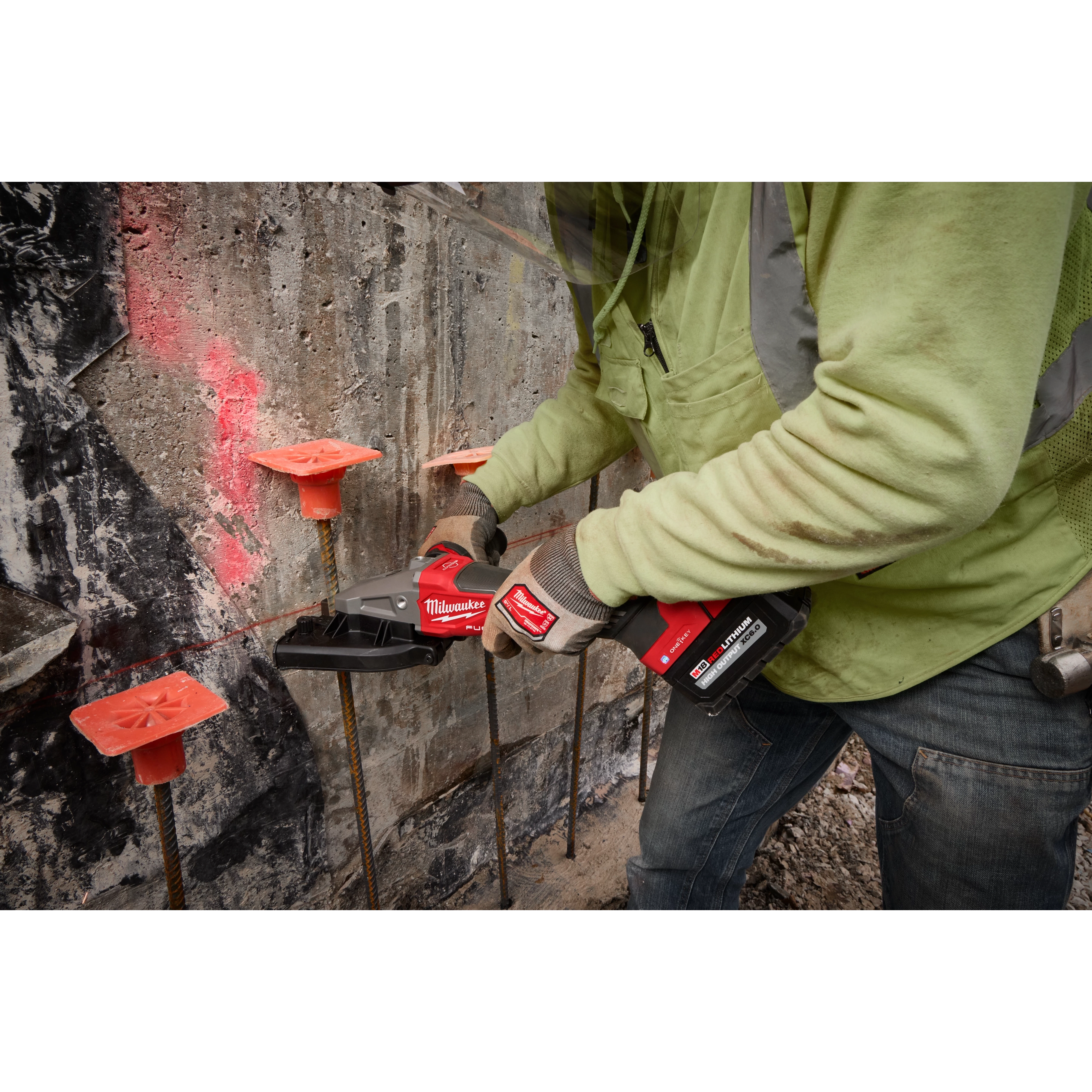 A person wearing a neon green jacket and gloves is using an M18 FUEL™ #10 (1-1/4") Rebar Cutter to cut through rebar protruding from a concrete wall. The rebar segments are topped with orange plastic caps for safety.