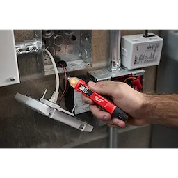 A person uses a Dual Range Voltage Detector to test wiring in an open electrical box. The red tool has a black tip and a glowing light, indicating the presence of voltage.