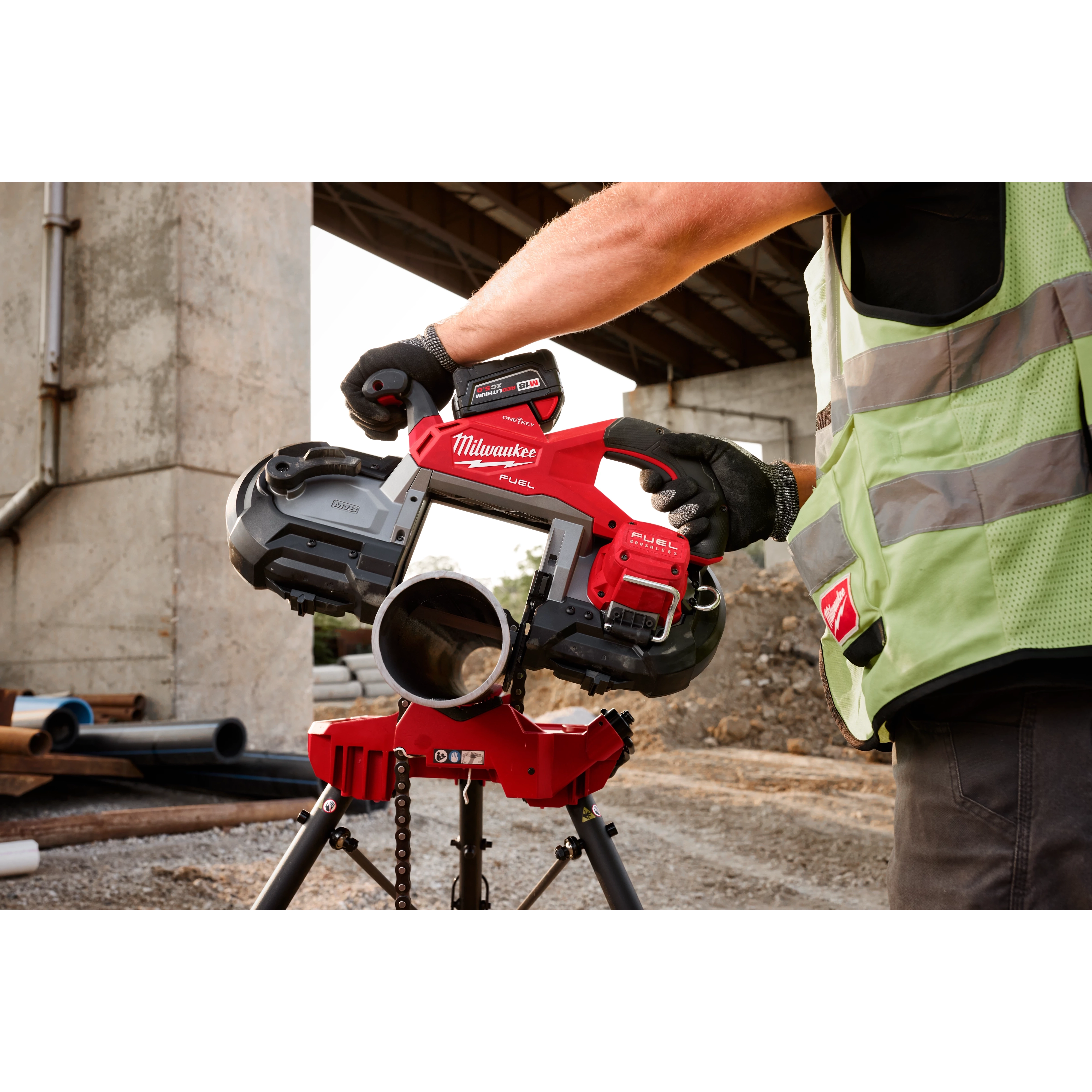 A person wearing gloves and a safety vest operates the M18 FUEL™ Deep Cut Dual Trigger Band Saw w/ ONE-KEY™ to cut a pipe on a construction site. The tool features a red and black color scheme and is mounted on a stand.