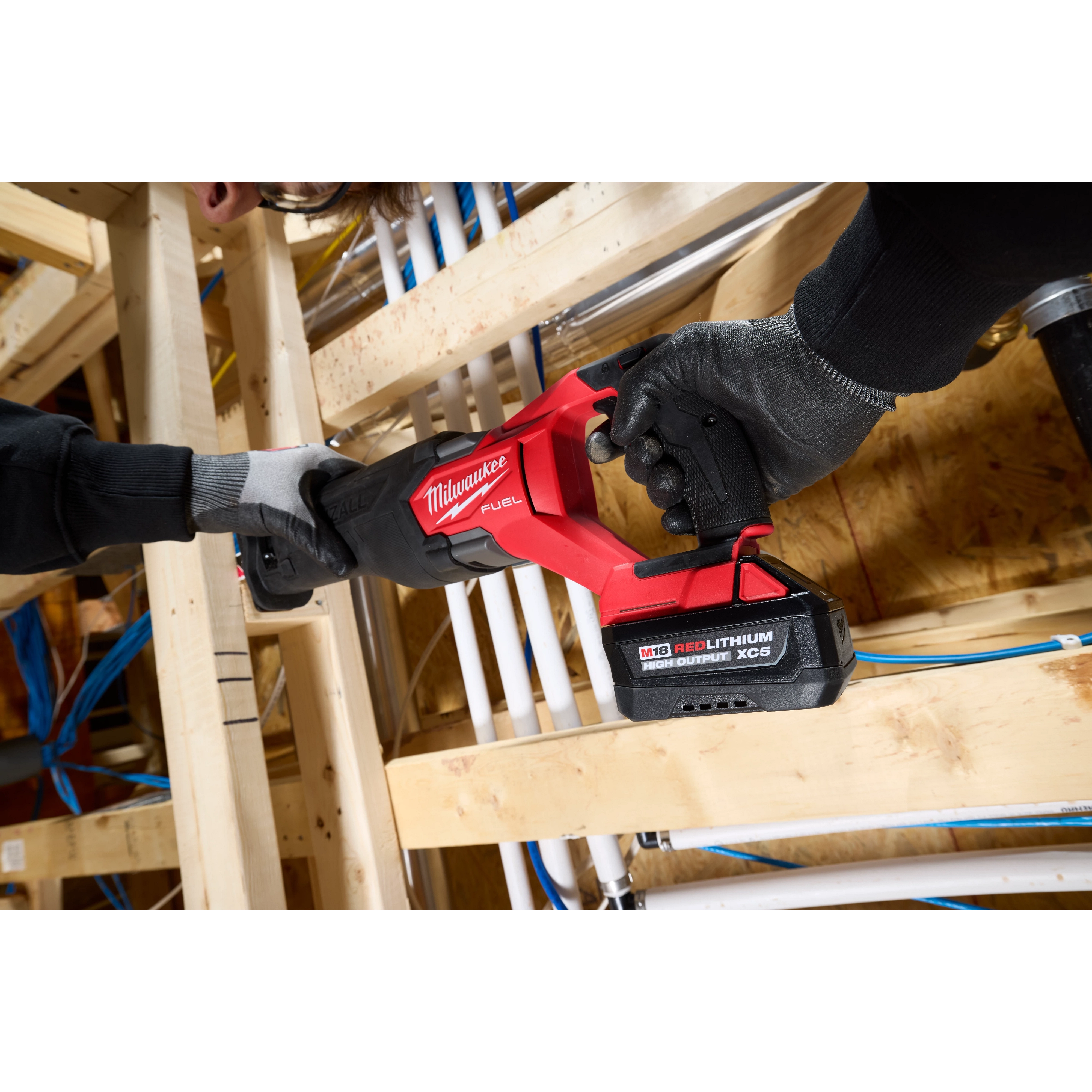 Worker using a red power saw with an M18 REDLITHIUM HIGH OUTPUT XC5 Battery Pack among wooden framing and pipes.