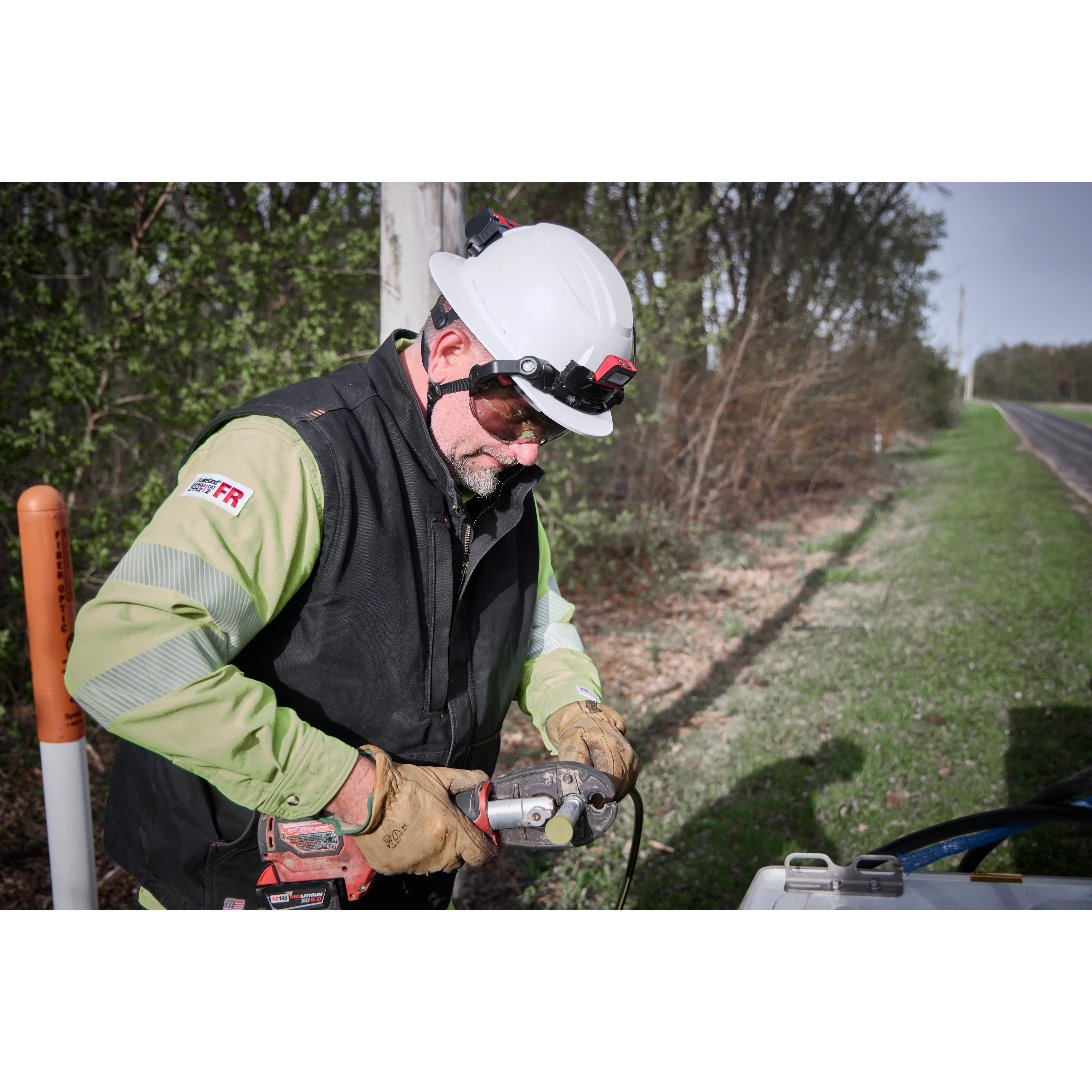A worker wearing a BOLT™ White Full Brim Safety Helmet – Type 2, Class E, uses an electric drill outdoors. The worker is dressed in a green and black jacket, brown gloves, and is standing next to a vehicle on the side of a rural road with trees in the background.