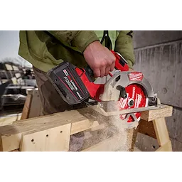 A person is using a power saw equipped with an M18™ REDLITHIUM™ FORGE™ HD12.0 Battery Pack to cut through a piece of wood. Dust and wood chips are seen from the cutting. The tool is being held firmly, and the scene appears to be an outdoor construction site.