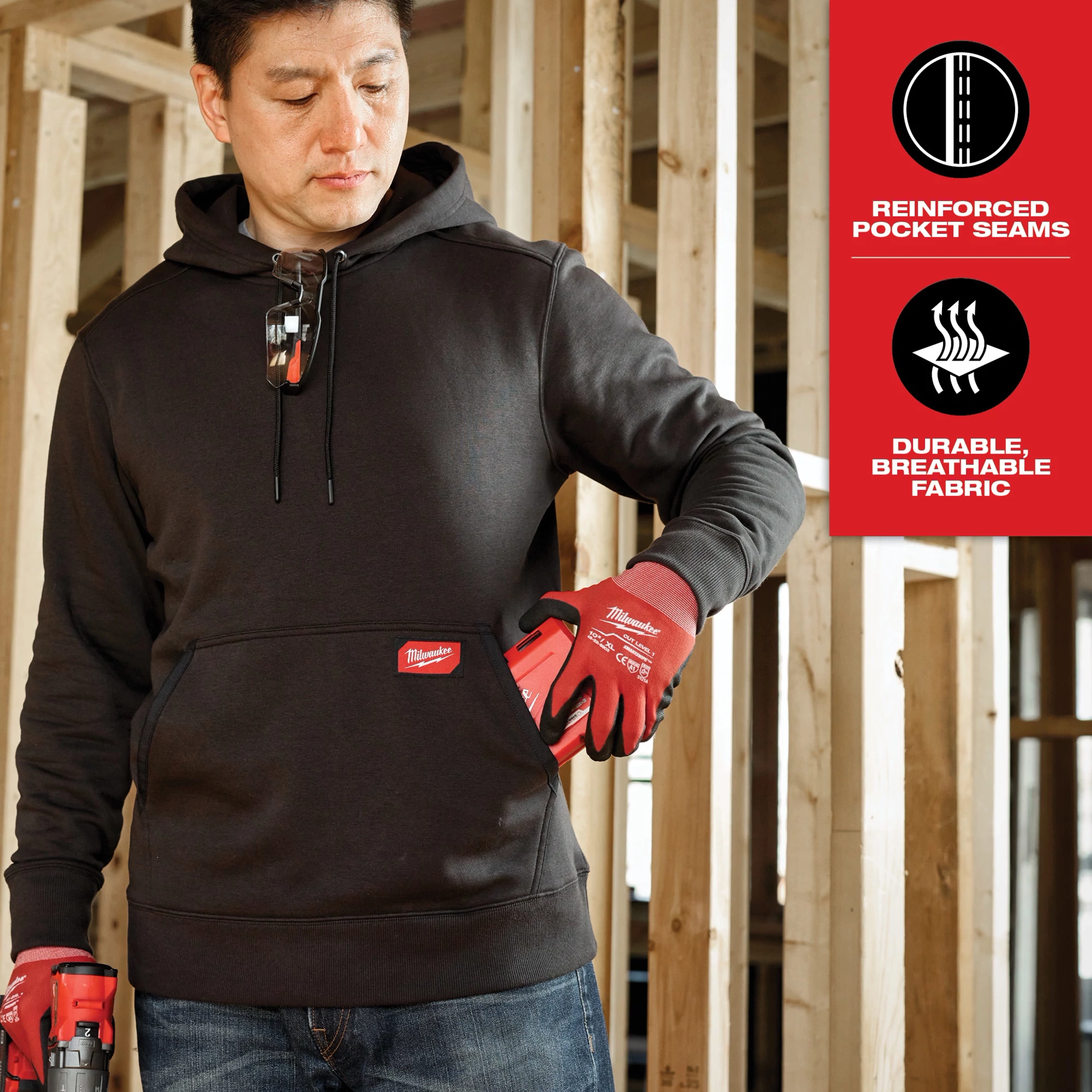 Man wearing black Midweight Pullover Hoodie standing in an unfinished construction setting, holding a red tool and wearing red gloves. Features reinforced pocket seams and breathable fabric.
