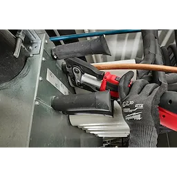 The ZoomLock MAX® & MaxiPro® Pivoting Press Ring Kit for M18™ FORCE LOGIC™ Press Tool is being used in a mechanical setting. A gloved hand operates the red press tool, handling pipes connected to a metal surface with labeled tags and several black and blue features for pipe fittings visible.