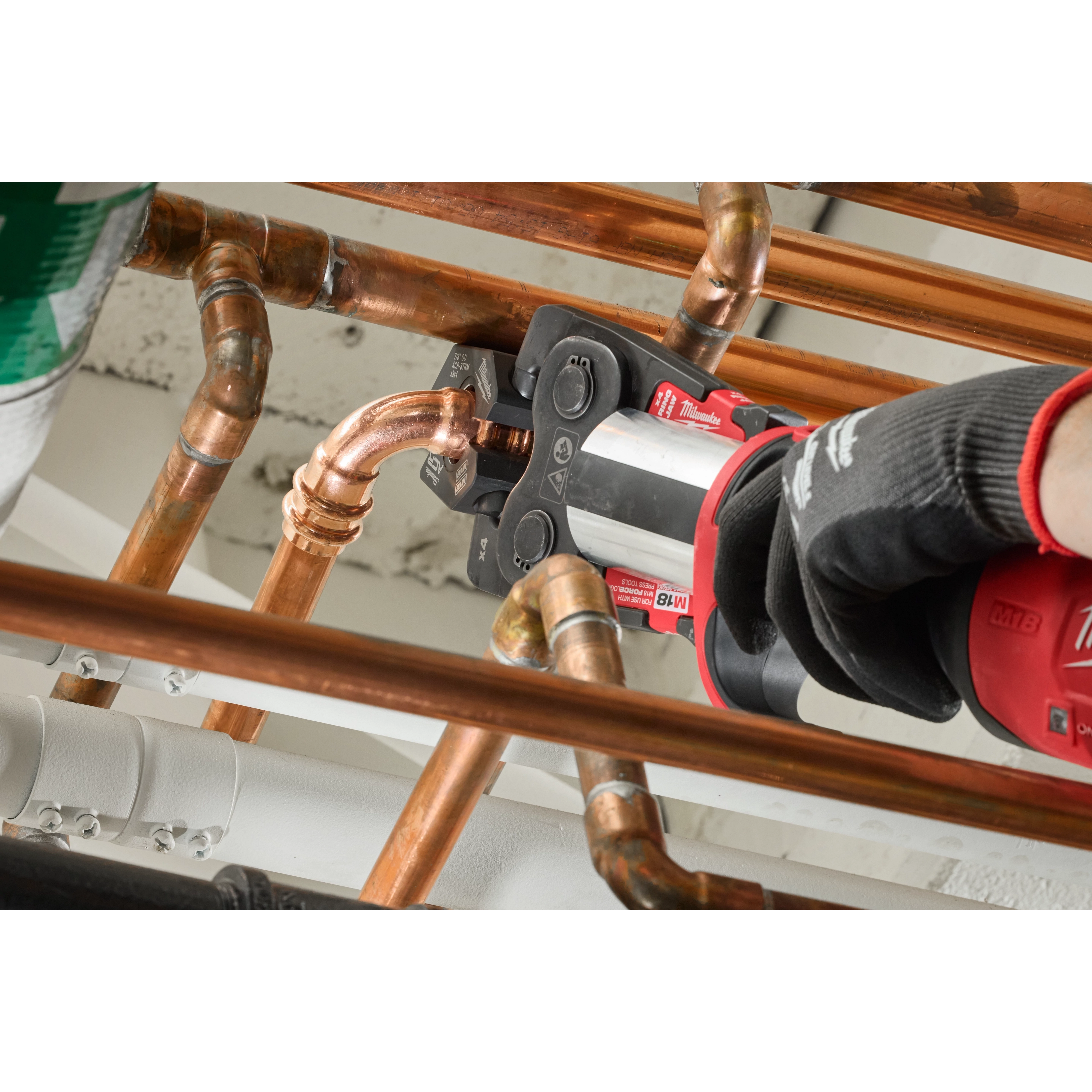 A gloved hand operates an M18™ FORCE LOGIC™ Press Tool to secure a copper pipe fitting using the 1/4" - 7/8" Streamline® ACR Pivoting Press Ring Kit. The tool is photographed amidst a network of copper pipes, demonstrating the pivoting press ring’s function in a plumbing installation.