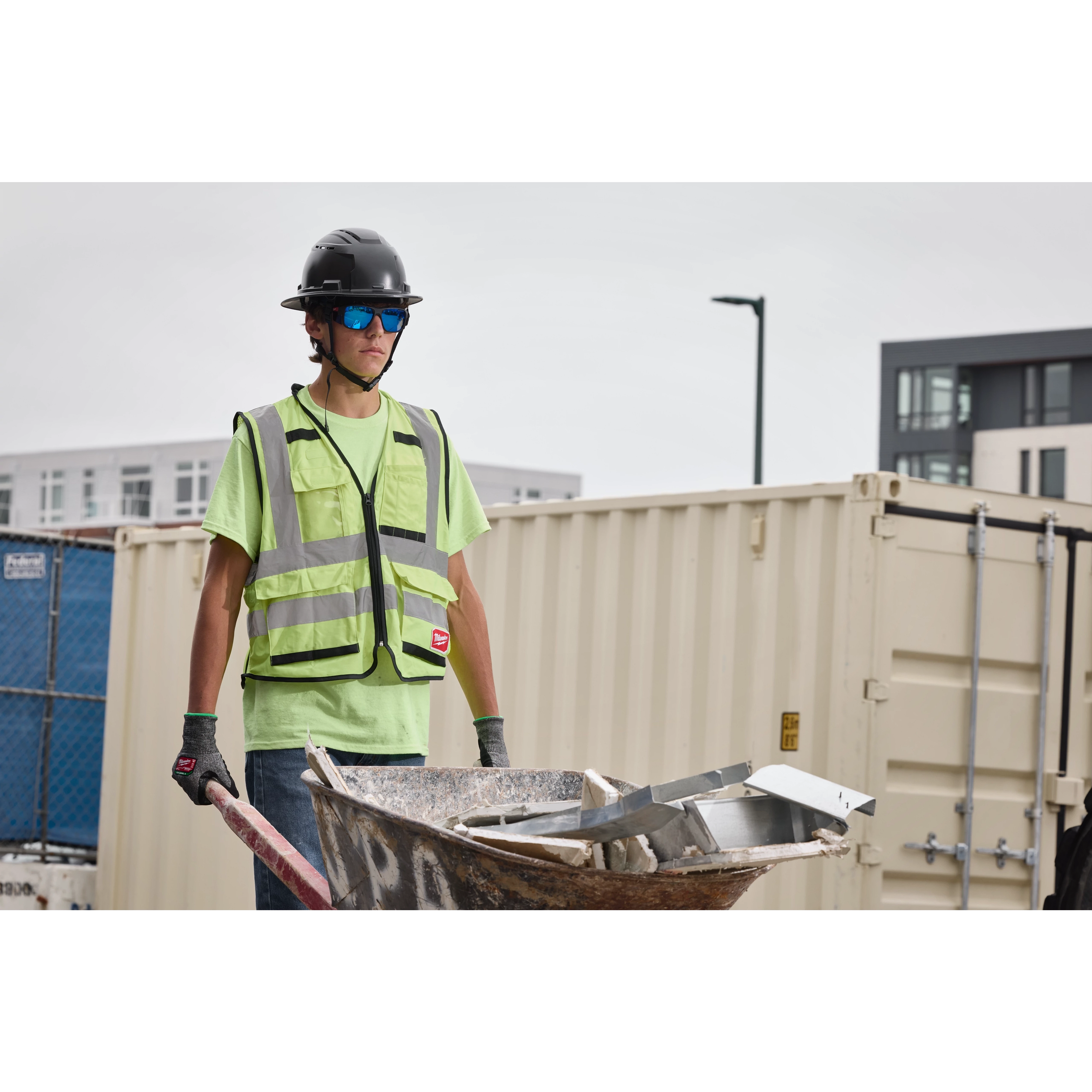 A construction worker wearing a green safety vest, helmet, and gloves holds a wheelbarrow filled with metal debris. The worker is wearing Full Frame Safety Glasses with Removable Side Shields – Blue Mirrored Anti-Scratch Lenses. The background includes shipping containers and buildings.