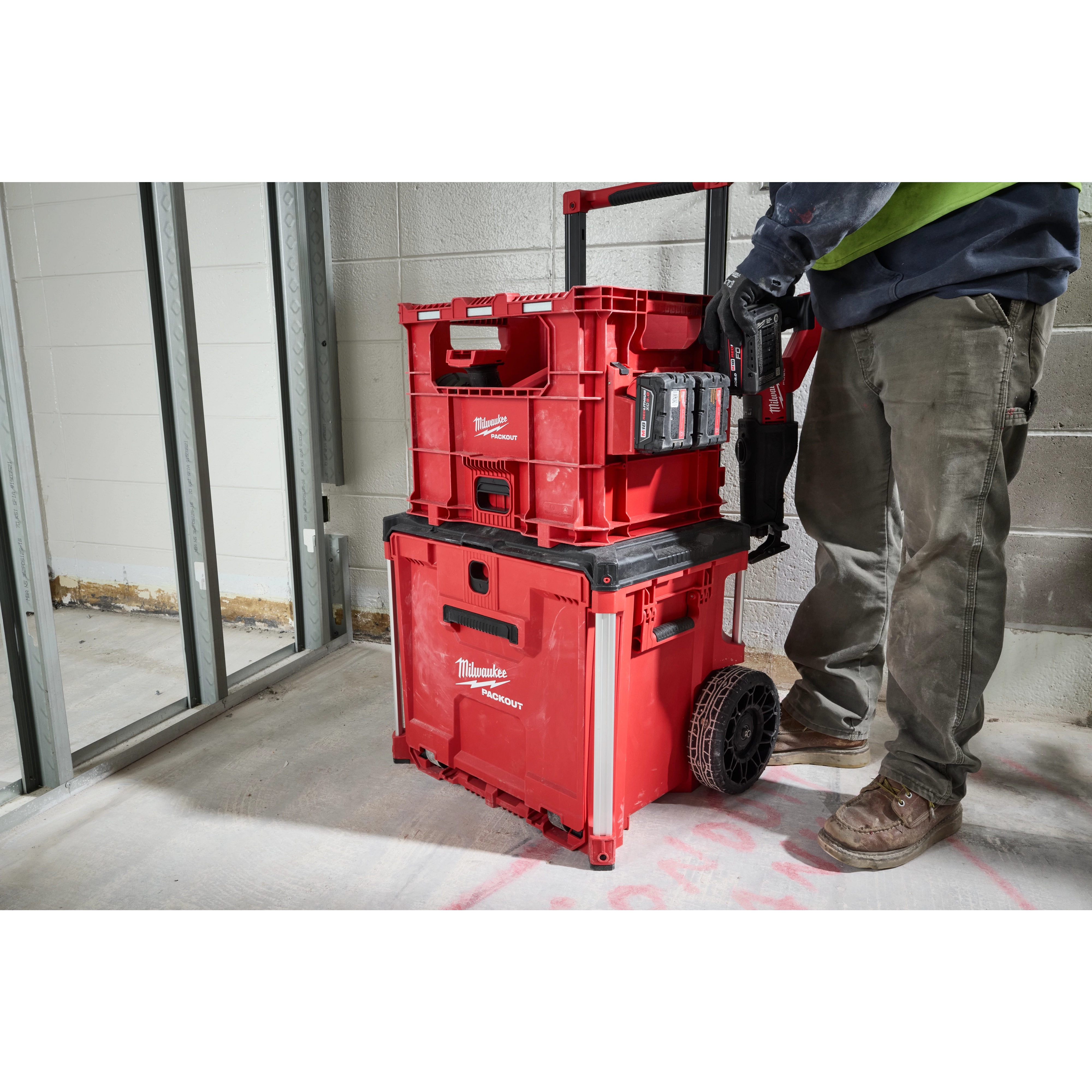 Person standing next to a stack of red PACKOUT XL Crate storage units, equipped with wheels and a handle, in a construction area.