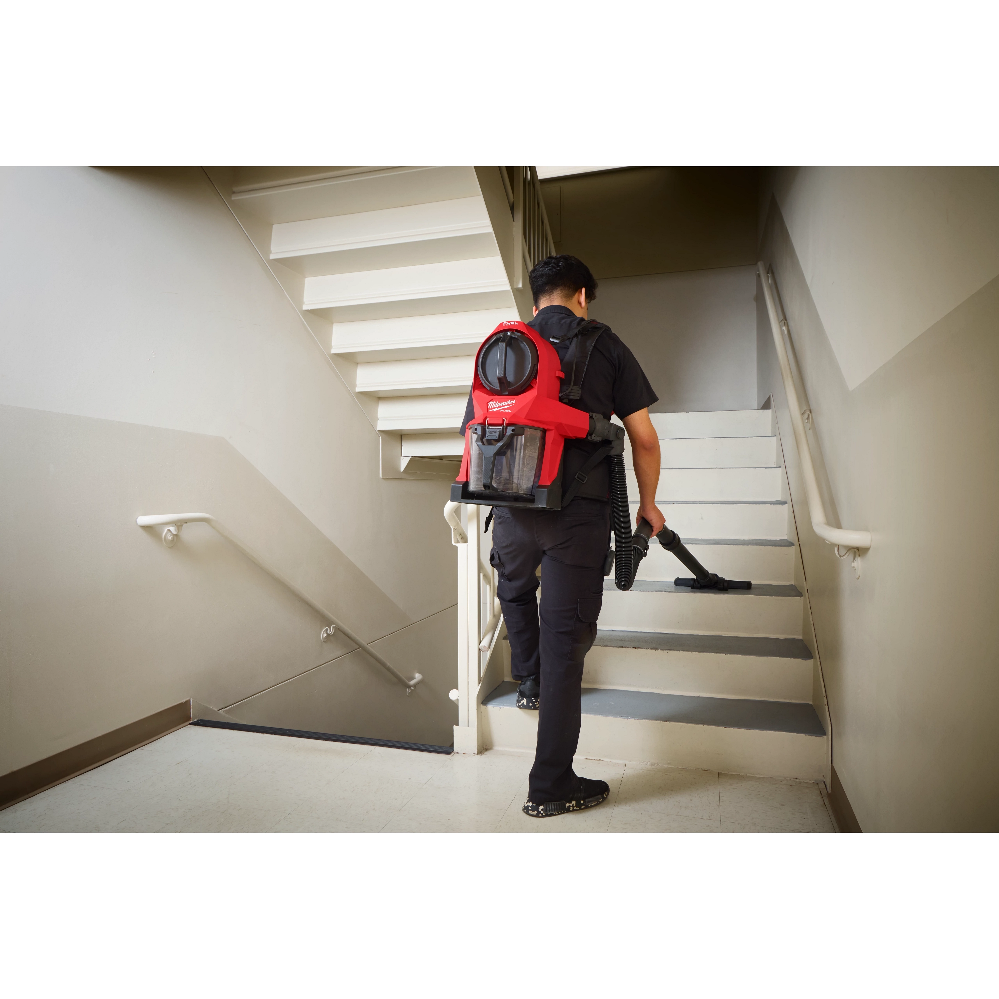 Person carrying and using the M18 FUEL™ 3-in-1 Backpack Vacuum while walking up a stairwell. The vacuum is red with black straps and has multiple compartments for different cleaning functions.