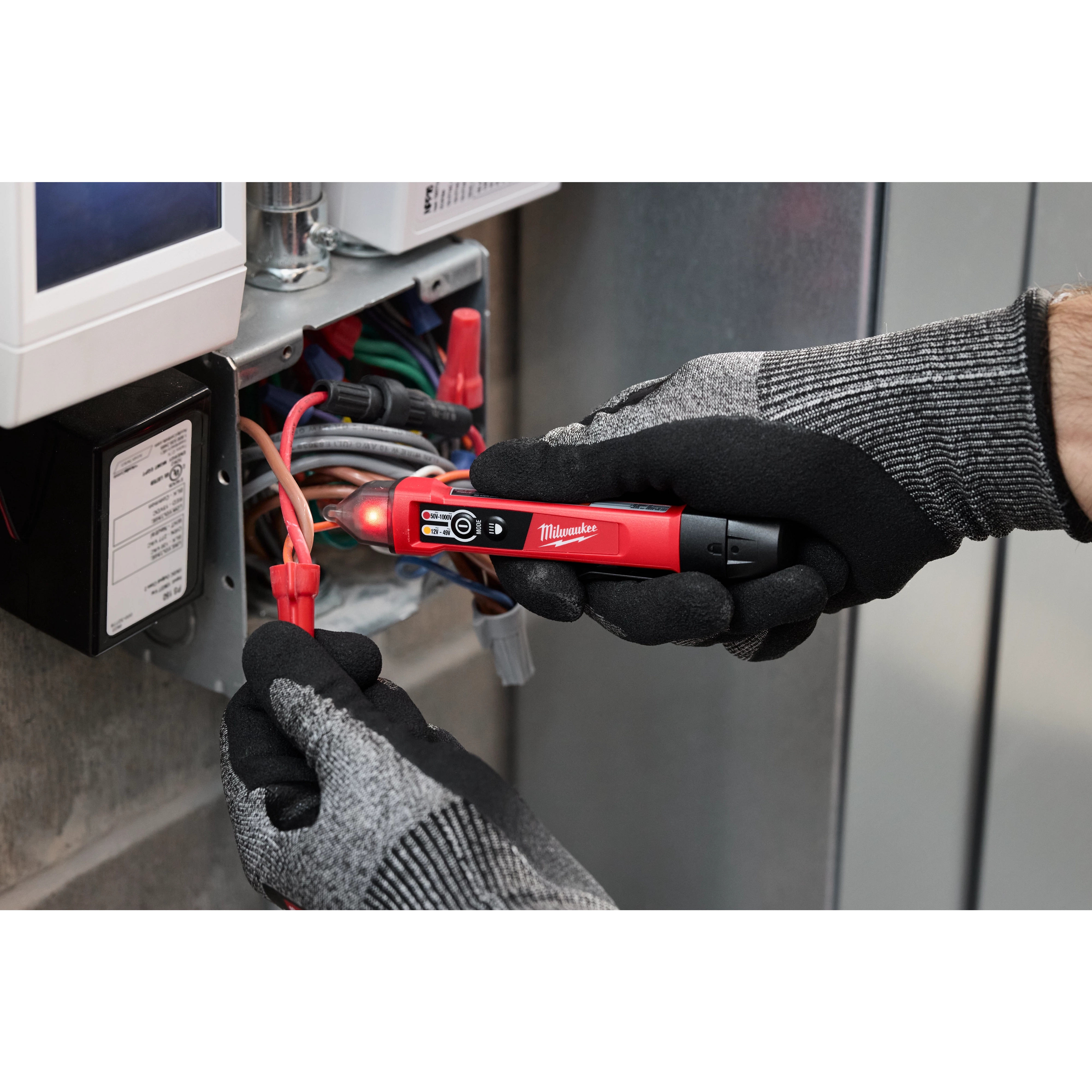 Person wearing gloves uses a Dual Range Voltage Detector w/ LED on a bundle of wires inside an electrical box. The tool's LED lights up, indicating voltage.