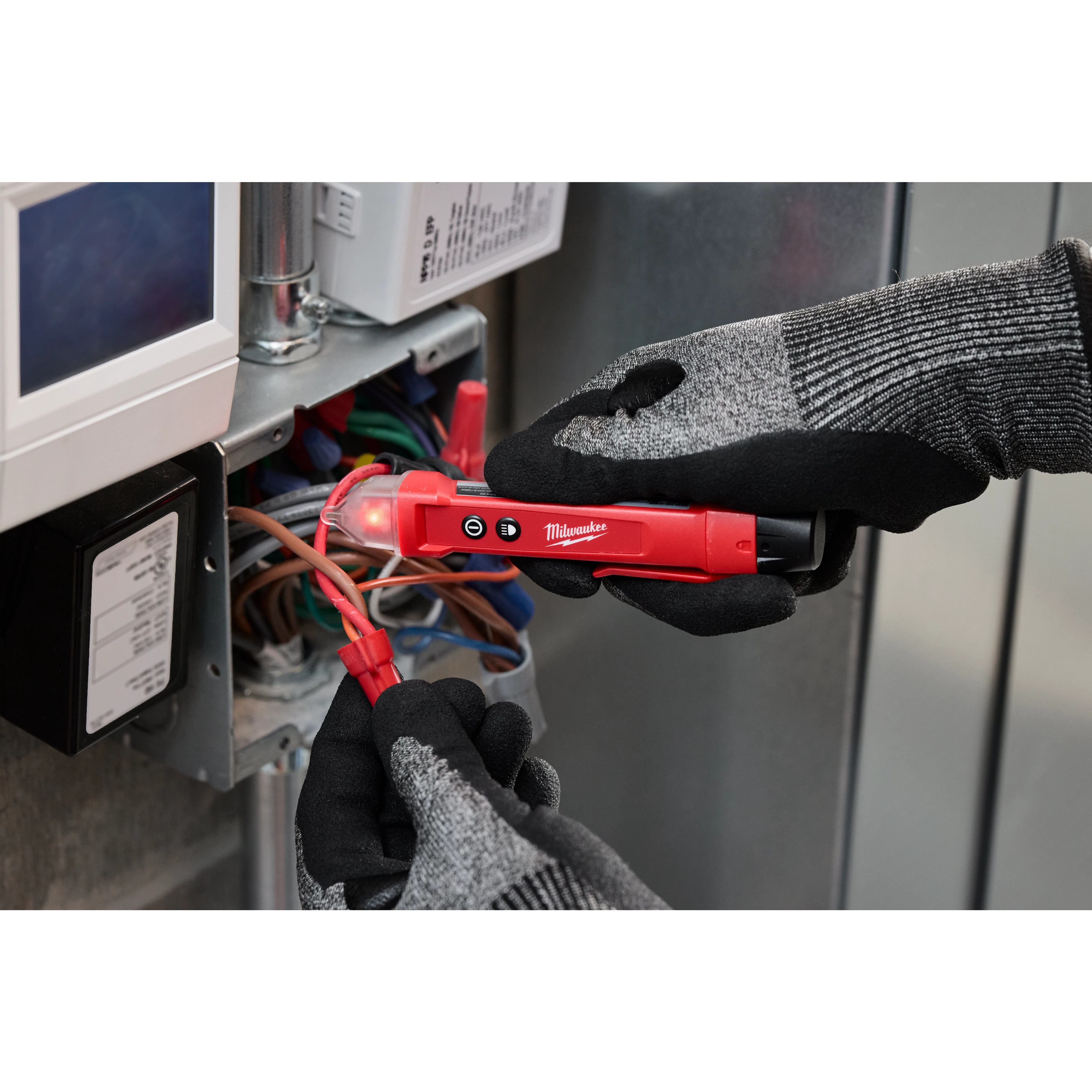 A person wearing black and gray gloves uses a red Voltage Detector w/ LED to check electrical wires inside an open electrical box.