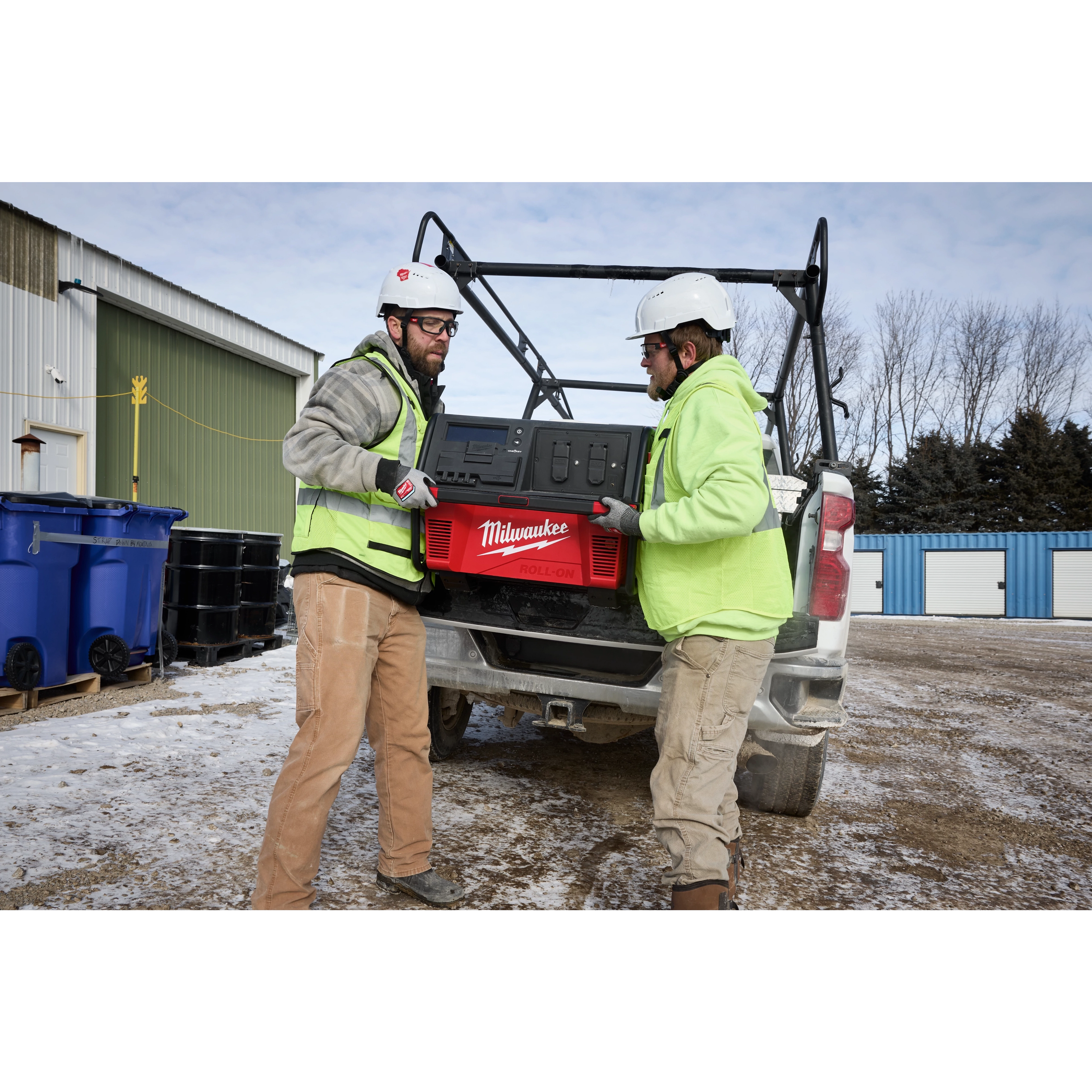 Two workers wearing helmets and uniforms lift a Milwaukee ROLL-ON 7200W/3600W 6.0kWh Power Supply into a pickup truck.