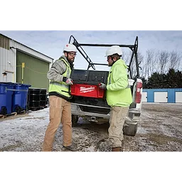 Two workers wearing helmets and uniforms lift a Milwaukee ROLL-ON 7200W/3600W 6.0kWh Power Supply into a pickup truck.