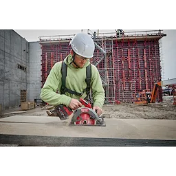 A construction worker uses the M18™ FUEL™ 7-1/4" Circular Saw w/ONEKEY™ to cut wood on a job site. The worker wears a green safety harness and operates the red and black saw that creates sawdust in the air. A large scaffold structure is built in the background.