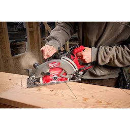 A person is using an M18 FUEL™ 7-1/4" Rear Handle Circular Saw to cut a piece of wood. The saw is red and black, and sawdust is visible in the air.