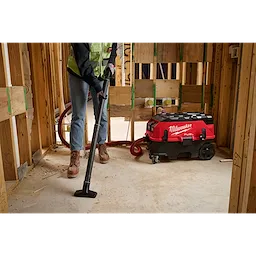 A person is using a vacuum from the PACKOUT™ Cleaning Accessory Kit to clean up a construction site. The vacuum is red and black and has "Milwaukee" written on it. The area is surrounded by unfinished wooden walls.