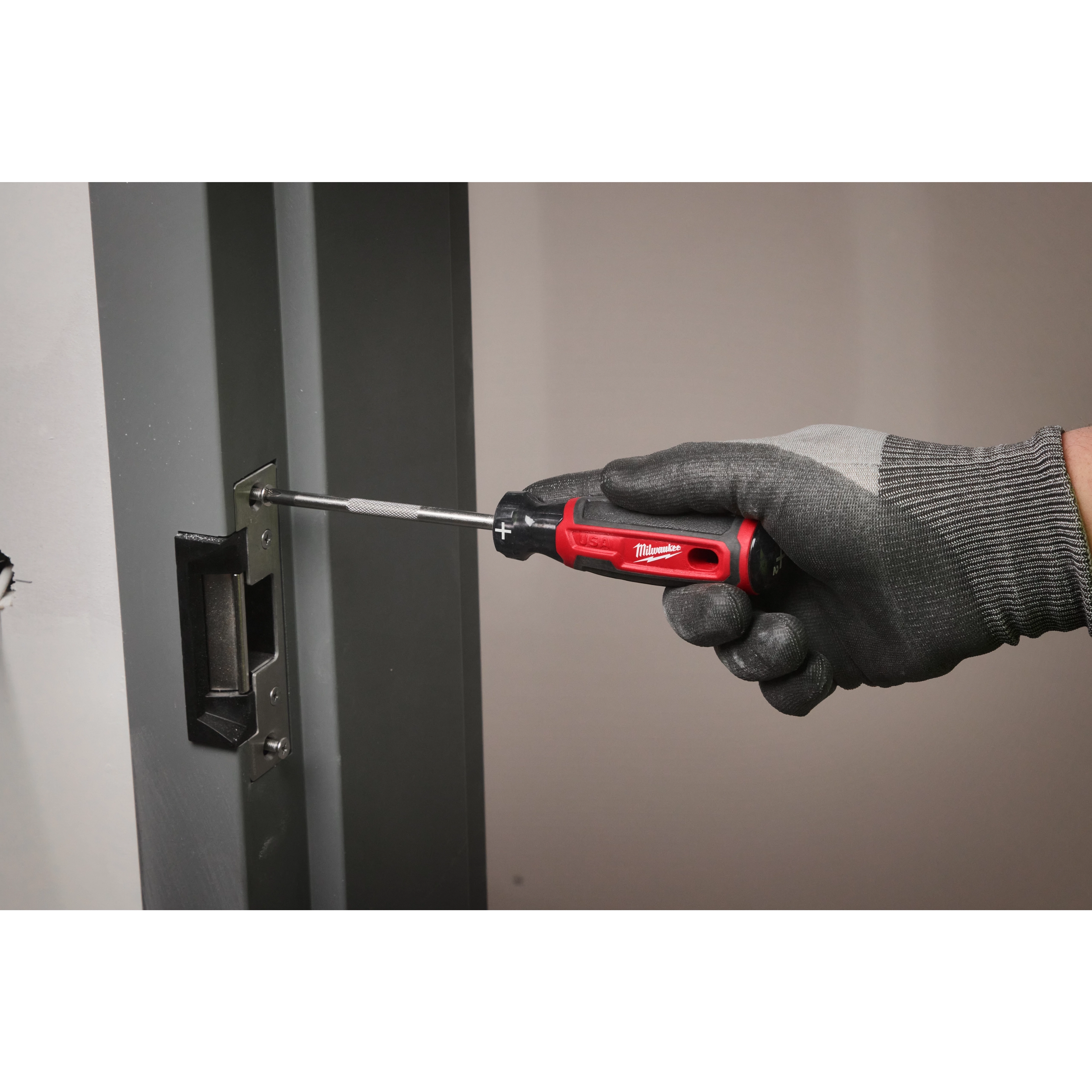 A gloved hand uses the #2 Phillips 4" Cushion Grip Screwdriver (USA) to tighten screws on a door lock mechanism. The screwdriver is red and black with a comfortable grip, ensuring ease of use during tasks.