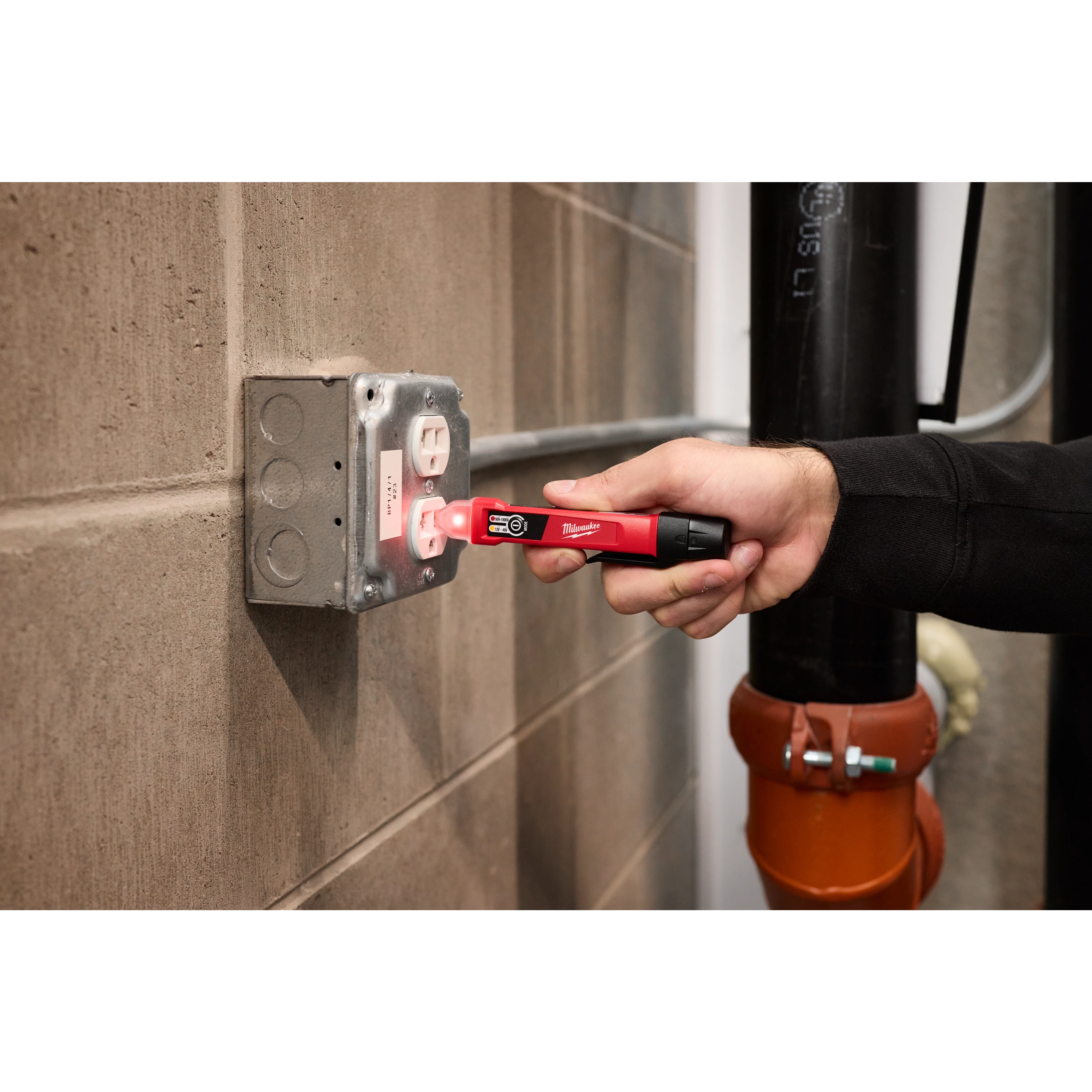 A person is using a Dual Range Voltage Detector to test an electrical outlet in a gray wall-mounted box.