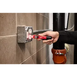 A person is using a Dual Range Voltage Detector to test an electrical outlet in a gray wall-mounted box.