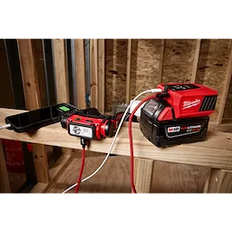 Milwaukee M18 REDLITHIUM XC5.0 battery powers a smartphone and a Milwaukee headlamp on a wooden beam within a construction site. The battery is connected via red and white cables, visible in an unfinished wood-framed room with raw insulation in the background.