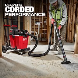 A worker uses the M18 FUEL™ NEXUS™ 12 Gallon Dual-Battery Wet/Dry Vacuum Kit to clean up construction debris. The red and black vacuum unit is equipped with wheels, a hose, and multiple battery slots. Text reads "Delivers Corded Performance." The background features an unfinished building interior.