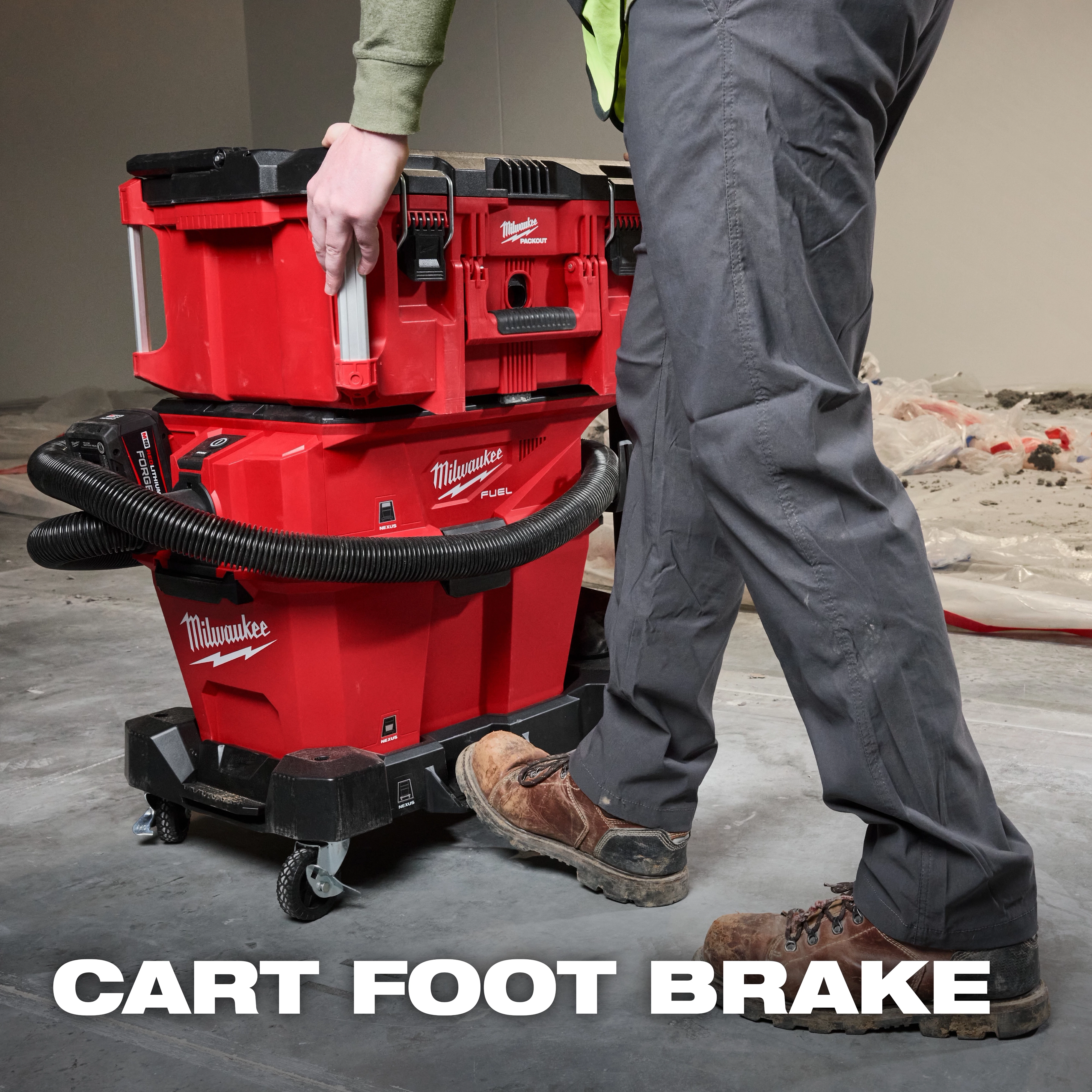 A person uses the foot brake on the Milwaukee NEXUS™ Wet/Dry Vacuum Cart. The cart is red and black, has a hose wrapped around it, and sits on caster wheels. The person's foot is pressing down on the brake pedal. Text reads "CART FOOT BRAKE."